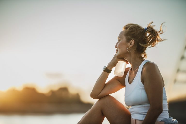 A middle age sportswoman sitting near the river and pensive looking into the distance while resting after training.