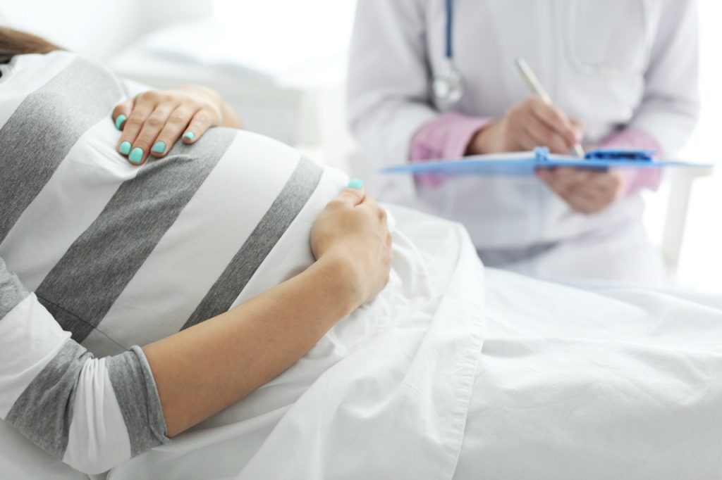 A pregnant woman lying down on a hospital bed while a healthcare provider in a white lab coat stands beside her, writing on a clipboard.