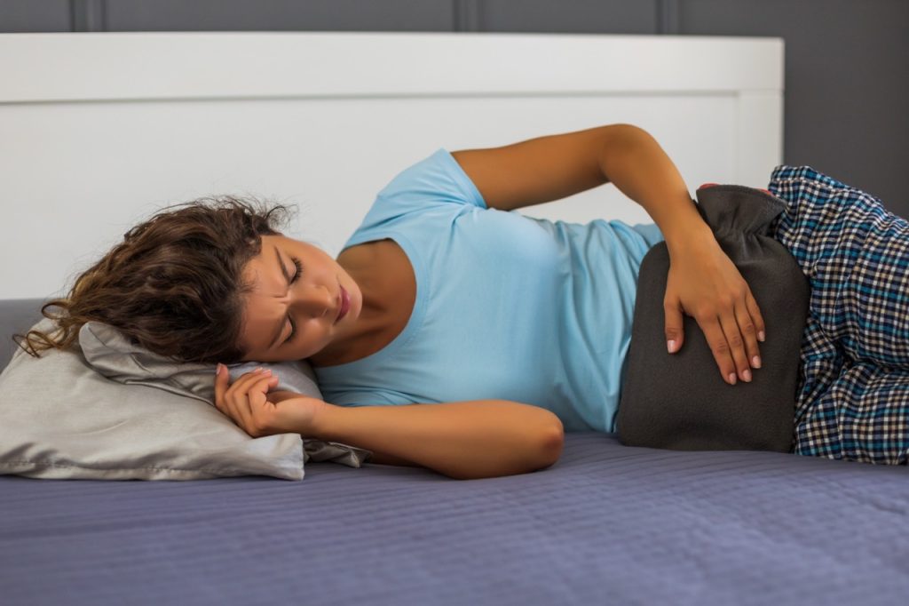 A woman lying on a couch holding a hot water bottle on her abdomen for pelvic pain relief.