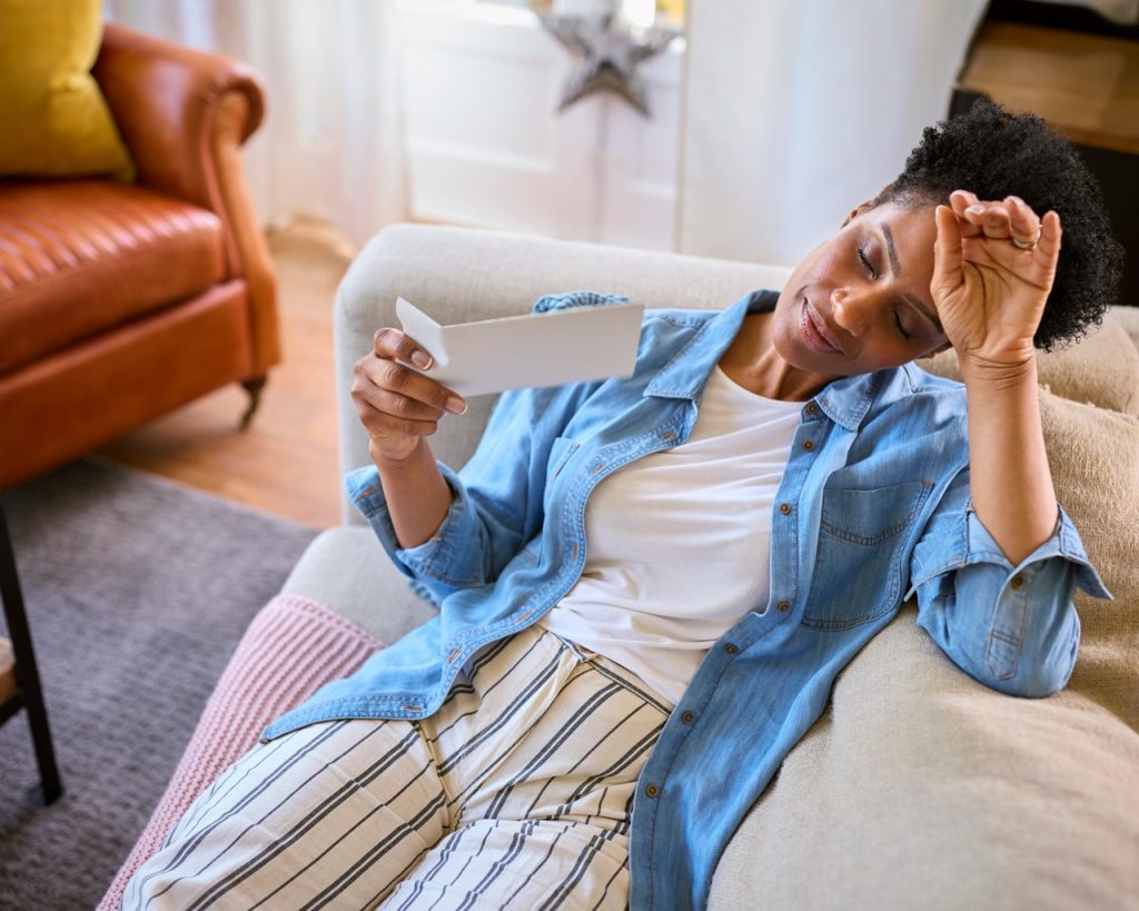 A mature woman sitting on a couch fanning herself with a hand fan to cool off from a hot flash during perimenopause.