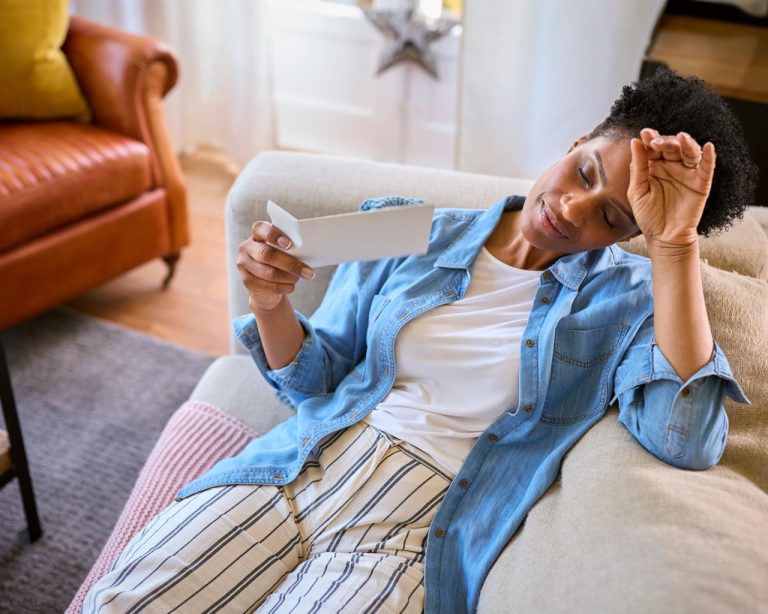 A mature woman sitting on a couch fanning herself with a hand fan to cool off from a hot flash during perimenopause.
