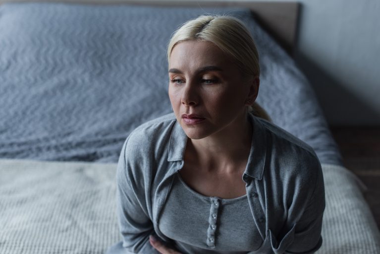A sad blonde woman sitting on a bed during menopause.